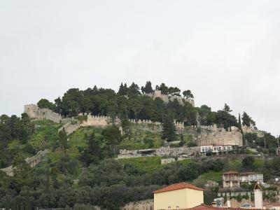 A stone fortress with crenellated walls and towers situated on a densely vegetated hill, overlooking a town with terracotta-roofed buildings in the foreground.