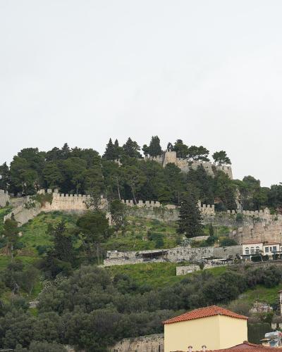 A stone fortress with crenellated walls and towers situated on a densely vegetated hill, overlooking a town with terracotta-roofed buildings in the foreground.