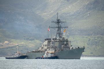 A large grey naval destroyer (hull number 66) is being assisted by two tugboats in a body of water with a steep, green, hilly coastline in the background.