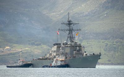 A large grey naval destroyer (hull number 66) is being assisted by two tugboats in a body of water with a steep, green, hilly coastline in the background.