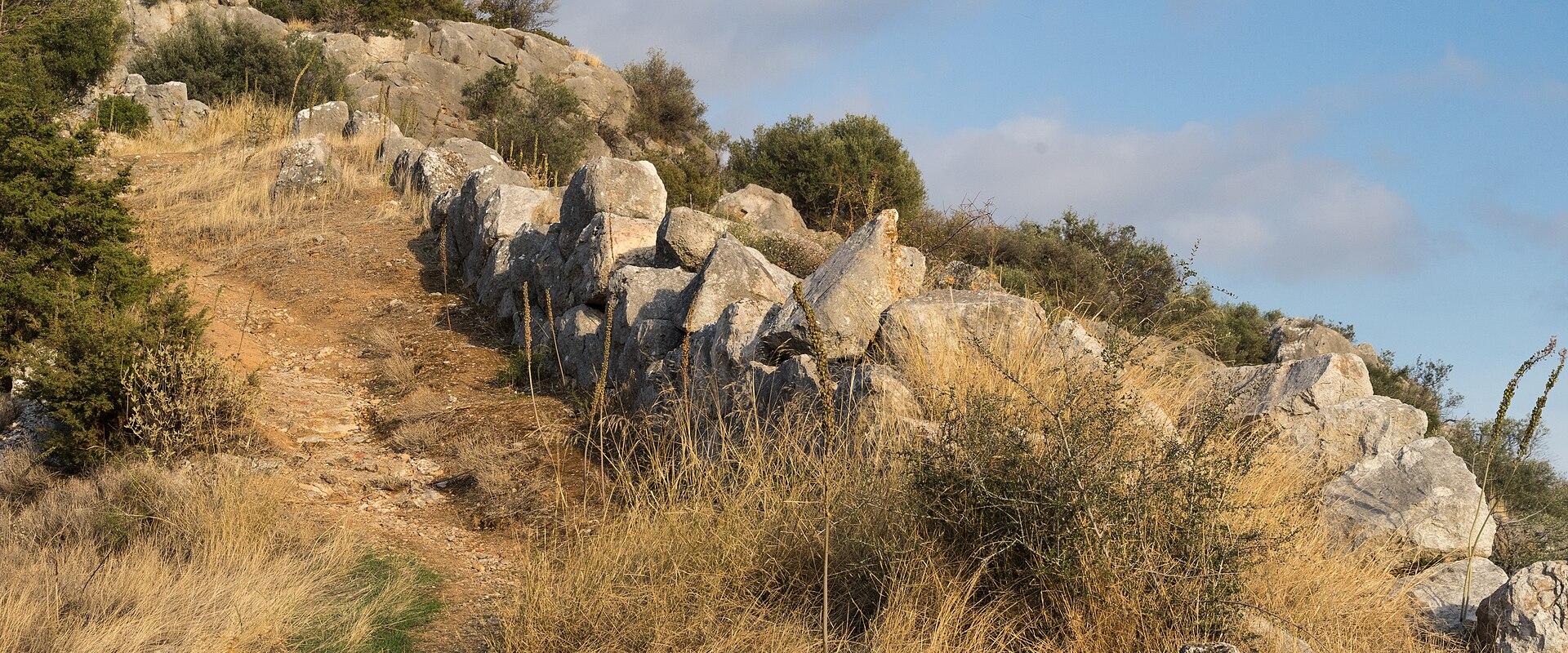 A low, dry-stone wall constructed of large, irregular rocks runs along a grassy, sunlit hillside under a blue sky.