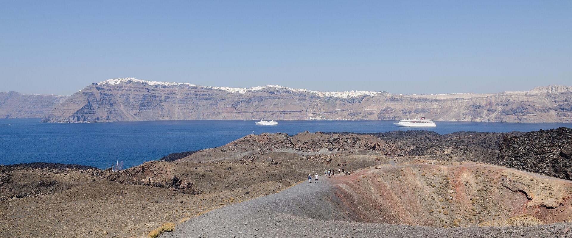 Aerial view of a barren, dark volcanic island surrounded by deep blue sea, with a large white cruise ship anchored nearby and other landmasses visible in the distance.