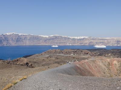 Aerial view of a barren, dark volcanic island surrounded by deep blue sea, with a large white cruise ship anchored nearby and other landmasses visible in the distance.