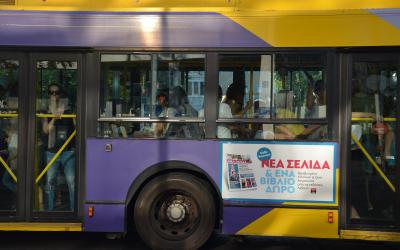 A side view of a purple and yellow public transit bus with passengers visible through the windows and an advertisement on the side panel.