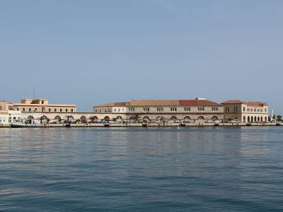 A waterfront view of a long, neoclassical building complex with arched colonnades along the quay, situated on a calm harbor with boats docked nearby.