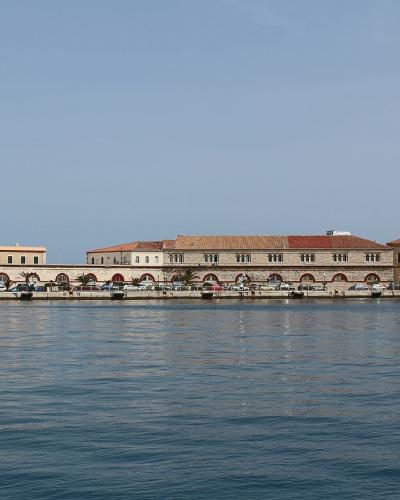 A waterfront view of a long, neoclassical building complex with arched colonnades along the quay, situated on a calm harbor with boats docked nearby.
