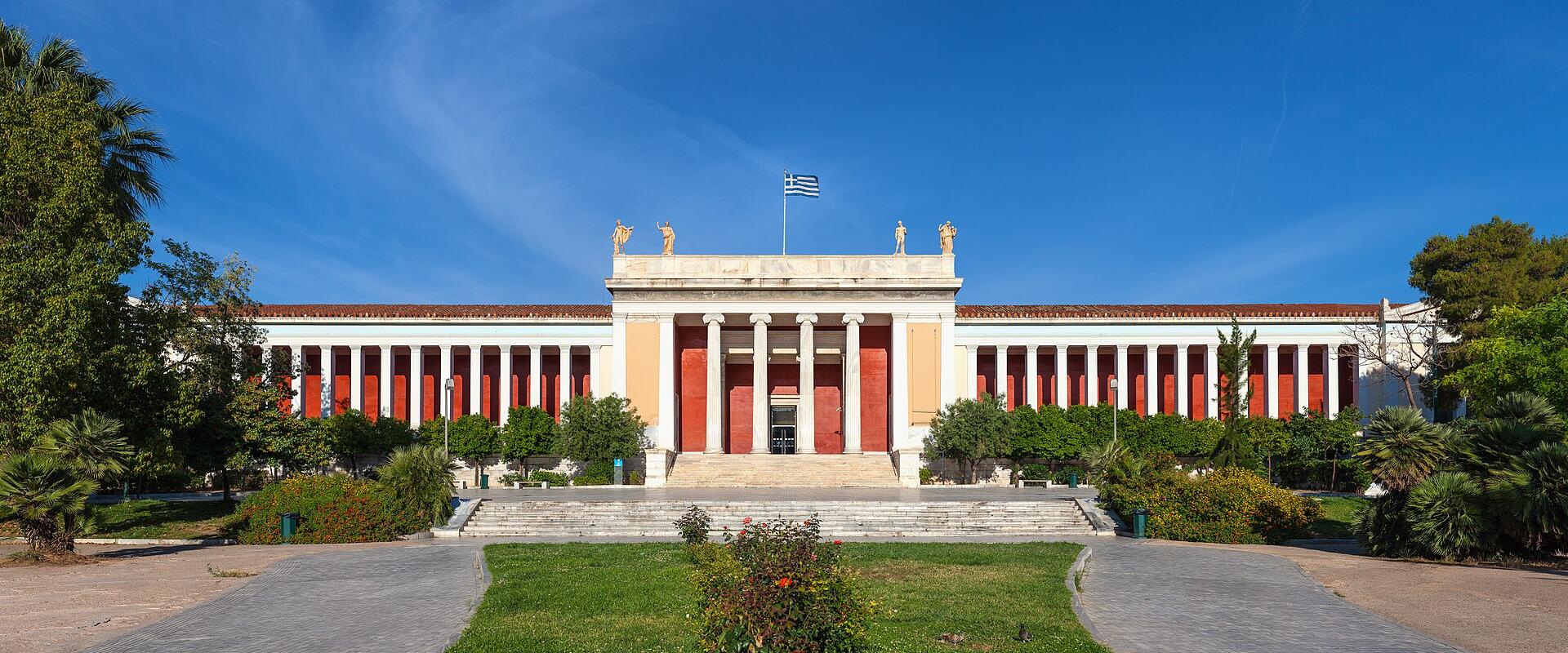 A large neoclassical building with a central portico, flanking colonnades, and a Greek flag flying above, set within a park-like garden.