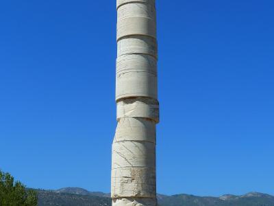 A tall, reconstructed ancient stone column standing on a stone base amidst ruins, with hills and a clear blue sky in the background.