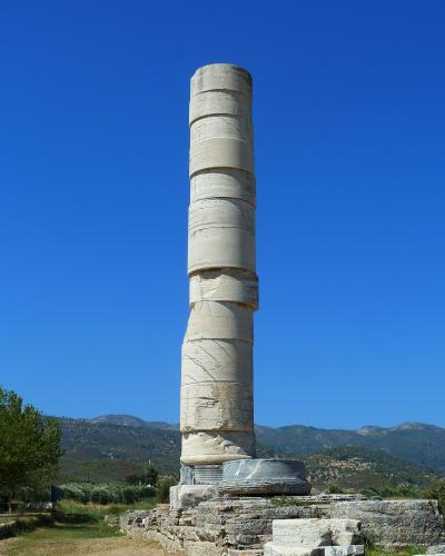 A tall, reconstructed ancient stone column standing on a stone base amidst ruins, with hills and a clear blue sky in the background.