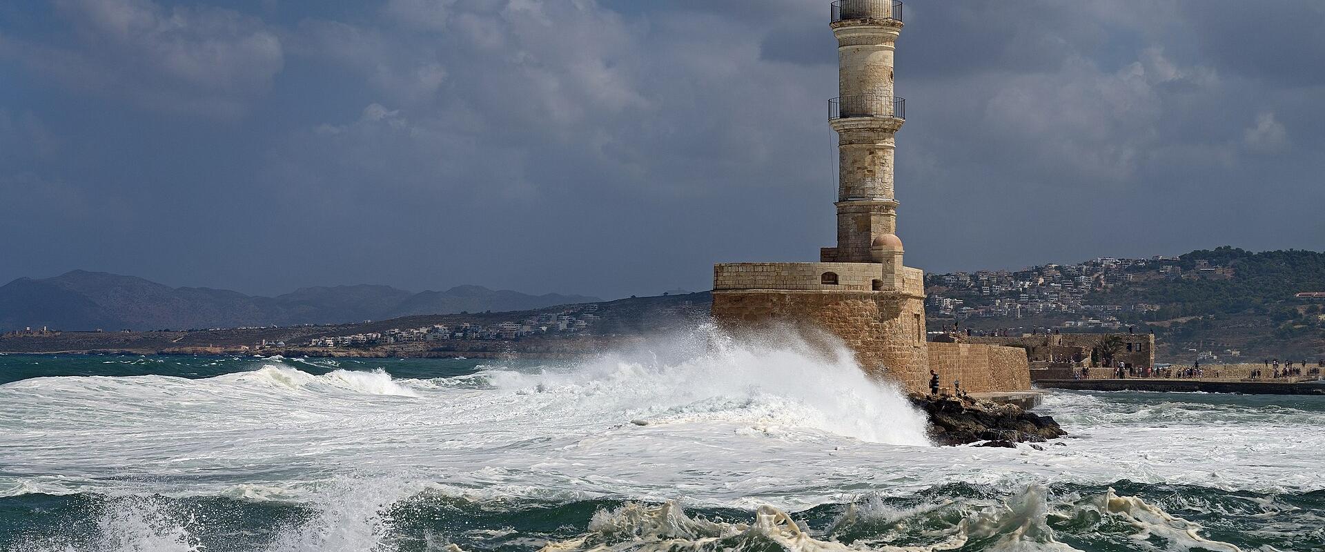 A tall, cylindrical stone lighthouse situated on a fortified breakwater, with large waves crashing against its base and a coastal town visible in the background under a cloudy sky.