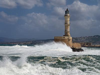 A tall, cylindrical stone lighthouse situated on a fortified breakwater, with large waves crashing against its base and a coastal town visible in the background under a cloudy sky.