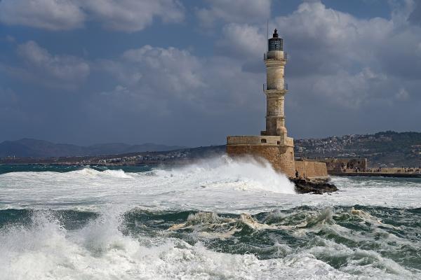 A tall, cylindrical stone lighthouse situated on a fortified breakwater, with large waves crashing against its base and a coastal town visible in the background under a cloudy sky.