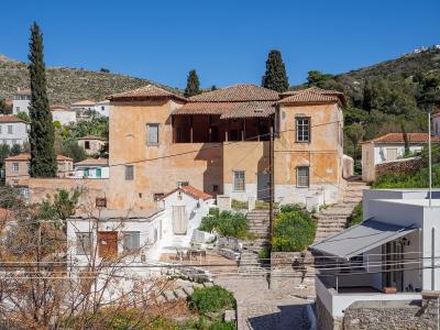 The image shows a large, traditional stone building with a terracotta tiled roof, situated in a hillside village setting, with a white building and a grey awning in the foreground.