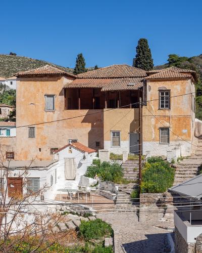 The image shows a large, traditional stone building with a terracotta tiled roof, situated in a hillside village setting, with a white building and a grey awning in the foreground.