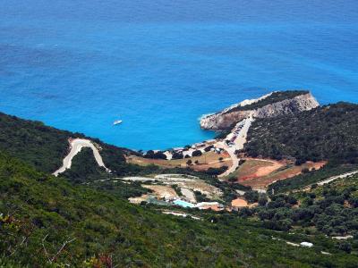 A high-angle landscape view of a coastal cove featuring a winding access road, a parking area with cars, a small building complex with a pool, and a distinctive white cliff promontory jutting into bright blue water.