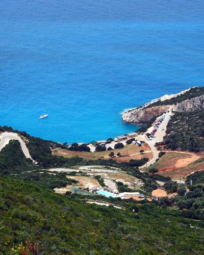A high-angle landscape view of a coastal cove featuring a winding access road, a parking area with cars, a small building complex with a pool, and a distinctive white cliff promontory jutting into bright blue water.