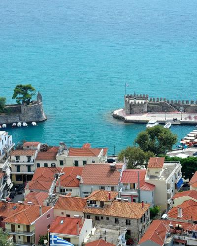 A high-angle view of a fortified harbor entrance with stone breakwaters, a small chapel on a rocky islet, and a dense cluster of buildings with red-tiled roofs in the foreground.