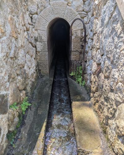 A stone-walled tunnel entrance with an arched opening, a metal gate swung open to the right, and a narrow concrete channel carrying flowing water leading into the dark tunnel.