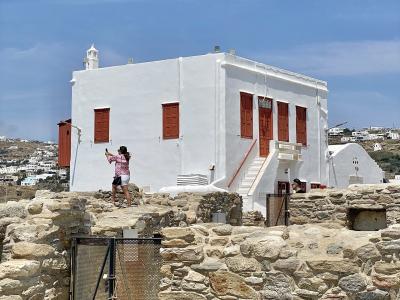 A white-washed building with reddish-brown wooden shutters and an external staircase, situated behind ancient stone ruins with a metal gate in the foreground.