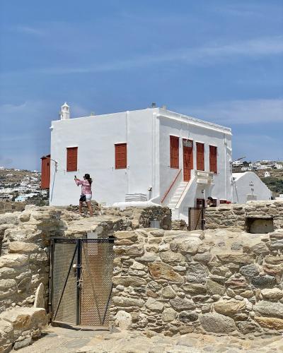 A white-washed building with reddish-brown wooden shutters and an external staircase, situated behind ancient stone ruins with a metal gate in the foreground.