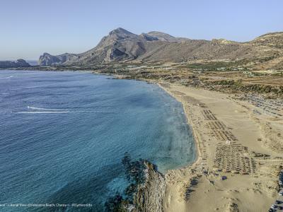 An aerial photograph of a long, sandy beach with rows of umbrellas and sunbeds, backed by arid hills and mountains, with a large rocky islet visible in the sea to the left.