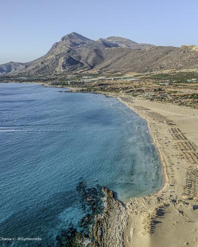 An aerial photograph of a long, sandy beach with rows of umbrellas and sunbeds, backed by arid hills and mountains, with a large rocky islet visible in the sea to the left.