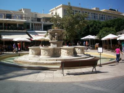 A stone fountain with four lions at the base and a central pedestal, situated in a paved public square surrounded by cafes and buildings.