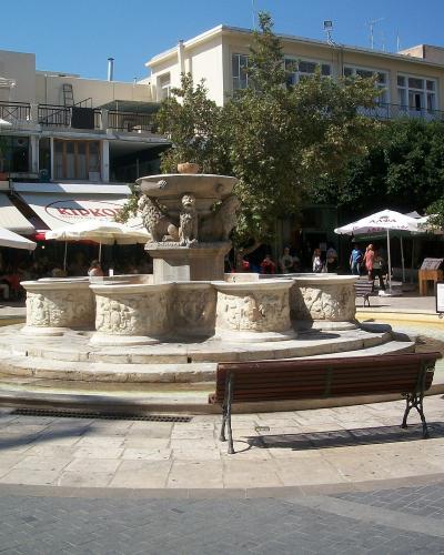 A stone fountain with four lions at the base and a central pedestal, situated in a paved public square surrounded by cafes and buildings.