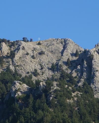 A rocky mountain ridge with scattered pine trees and a white cross visible at the summit against a clear blue sky.