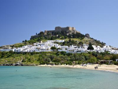 A coastal scene showing a sandy beach with turquoise water in the foreground, a hillside village of white cubic buildings in the middle ground, and a large ancient fortress perched on the summit of the hill.