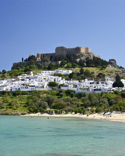 A coastal scene showing a sandy beach with turquoise water in the foreground, a hillside village of white cubic buildings in the middle ground, and a large ancient fortress perched on the summit of the hill.