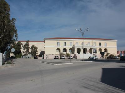 A two-story beige building with a red-tiled roof, arched colonnades on the ground floor, and a paved parking area in front, situated under a blue sky with scattered clouds.
