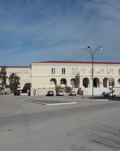 A two-story beige building with a red-tiled roof, arched colonnades on the ground floor, and a paved parking area in front, situated under a blue sky with scattered clouds.