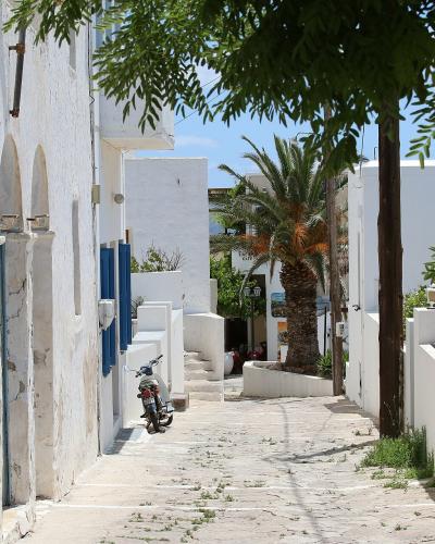 A narrow, paved street in a traditional white-washed village with blue shutters, a parked scooter, and a palm tree.