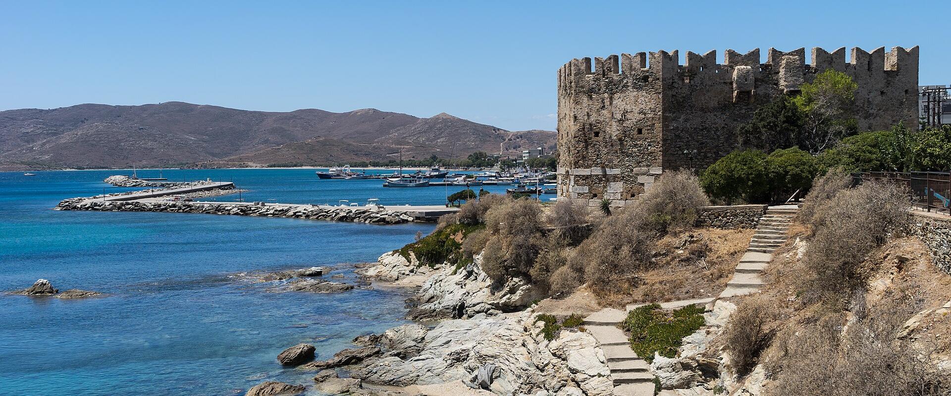 A stone fortress with crenellated walls stands on a rocky promontory overlooking a harbor filled with boats, with a small sandy beach and stone steps in the foreground.