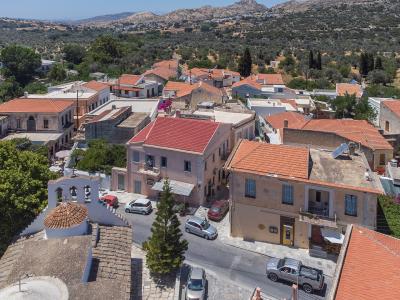 An elevated view of a Greek village with terracotta-roofed buildings, a white church with a dome and bell tower in the foreground, and a backdrop of olive groves and hills.