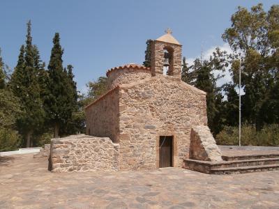 A small, rustic stone church with a bell tower and cross, situated in a paved courtyard surrounded by trees under a blue sky.
