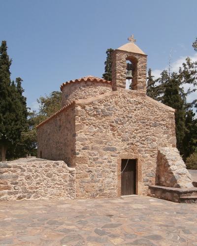 A small, rustic stone church with a bell tower and cross, situated in a paved courtyard surrounded by trees under a blue sky.