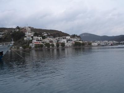 A coastal view of a Greek island town with white buildings climbing a hillside, a boat docked in the foreground, and a distant hill with a communications tower.