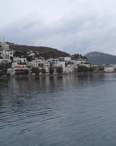 A coastal view of a Greek island town with white buildings climbing a hillside, a boat docked in the foreground, and a distant hill with a communications tower.