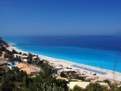A high-angle panoramic view of a long, sandy beach with turquoise water, lined with rows of blue and white sun umbrellas, adjacent to buildings with terracotta roofs and green vegetation.