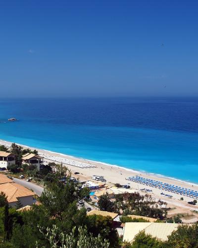 A high-angle panoramic view of a long, sandy beach with turquoise water, lined with rows of blue and white sun umbrellas, adjacent to buildings with terracotta roofs and green vegetation.