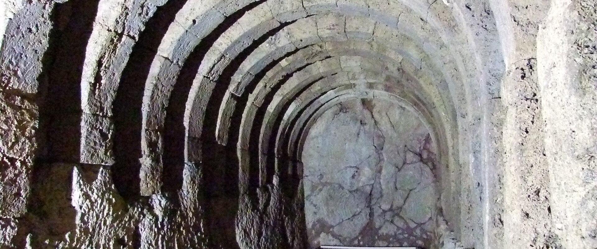 The image shows a stone-vaulted underground passage or tunnel with a metal handrail, likely part of an archaeological site's subterranean structure.