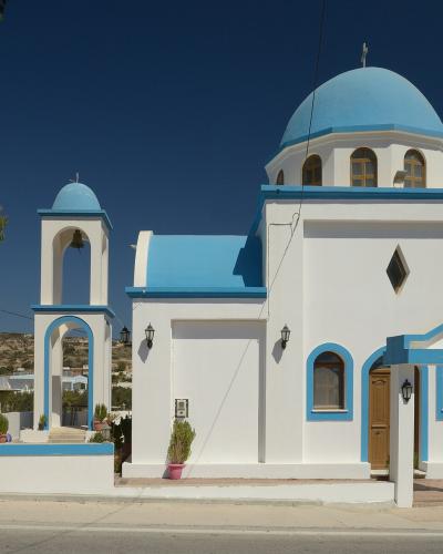 A white-washed Greek Orthodox church with bright blue domes and a bell tower, situated on a roadside with arid hills in the background.