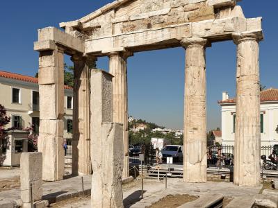 The image shows the Gate of Athena Archegetis, a stone monument with four columns and a pediment, located within an archaeological site with modern buildings visible in the background.