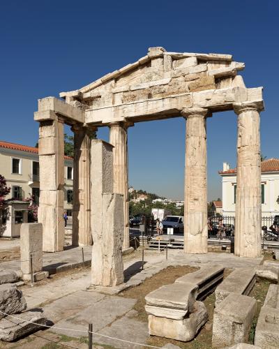 The image shows the Gate of Athena Archegetis, a stone monument with four columns and a pediment, located within an archaeological site with modern buildings visible in the background.