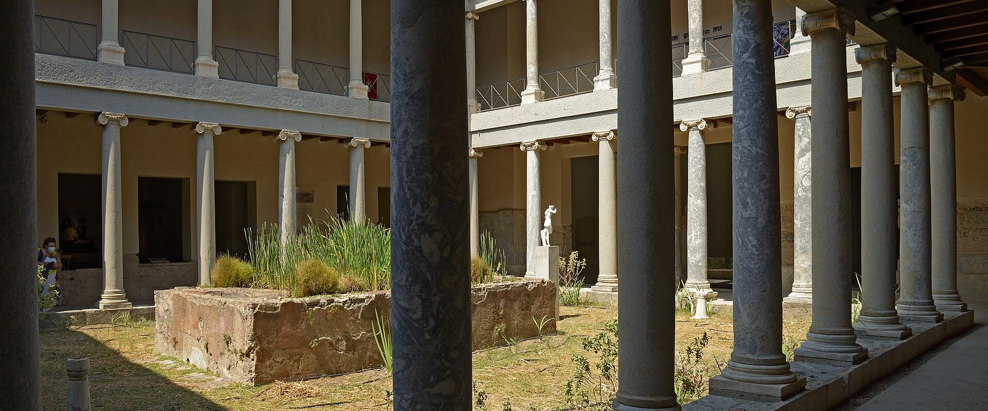 A view of a reconstructed Roman villa courtyard featuring a central garden bed, surrounded by a two-story colonnade with marble columns and a tiled roof.