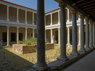 A view of a reconstructed Roman villa courtyard featuring a central garden bed, surrounded by a two-story colonnade with marble columns and a tiled roof.