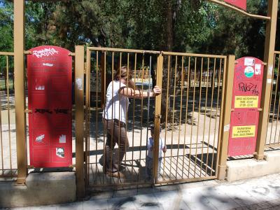 A woman and a child are entering a playground through a metal gate, with red informational signs on the gate posts displaying Greek text and symbols.