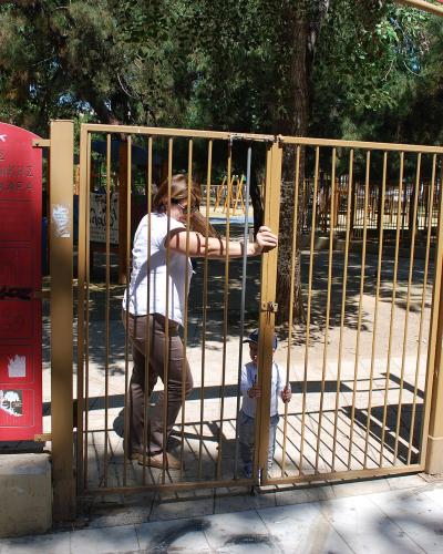 A woman and a child are entering a playground through a metal gate, with red informational signs on the gate posts displaying Greek text and symbols.